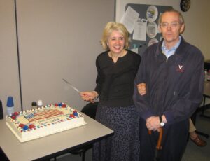A cake decorated with an American flag; beside it, a woman holding a bread knife stands arm-in-arm with a man leaning on an umbrella.