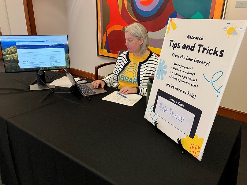 A librarian sits at a table in the main hallway of the law school. On one side of her is a computer screen; on the other side is a sign that reads "Research Tips and Tricks."